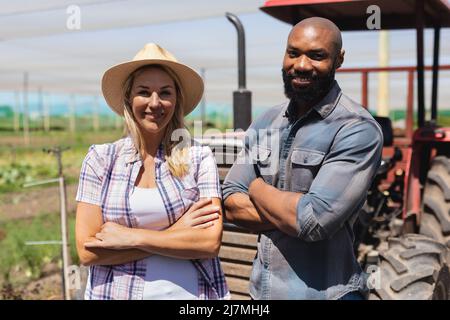 Smiling african american man and caucasian woman with arms crossed standing by tractor in greenhouse Stock Photo