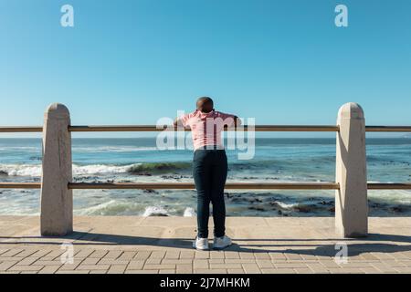 African american boy leaning on railing of promenade looking at sea against blue sky with copy space Stock Photo