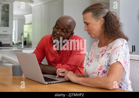 Multiracial senior couple using laptop while sitting at table in living room. unaltered, lifestyle, retirement, togetherness, wireless technology. Stock Photo