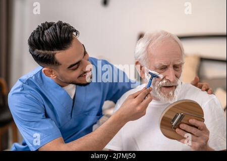 Joyous young man shaving a serious old gentleman Stock Photo