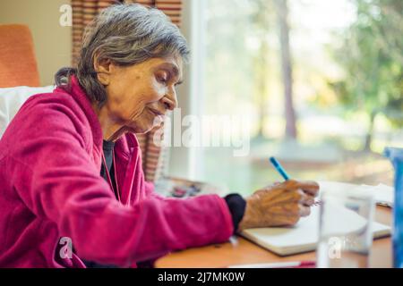Old elderly Asian Indian woman sitting writing in a journal or notebook, UK. Depicts memory loss and keeping a list or diary. Stock Photo