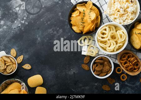 Unhealthy food. Snacks. All classic potato snacks with peanuts, popcorn and onion rings and salted pretzels in bowl plates on black concrete backgroun Stock Photo