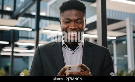 Happy smiling african guy american man businessman boss employee holding modern wireless device texting message on smartphone using social media apps Stock Photo