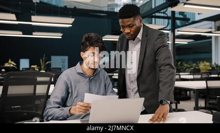 Male african mentor financial advisor businessman boss helping arab man colleague discussing marketing result in report paperwork american supervisor Stock Photo
