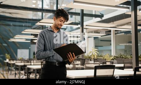 Young arabian confident business man guy stand in modern office hold folder check document male hispanic entrepreneur accountant read financial report Stock Photo