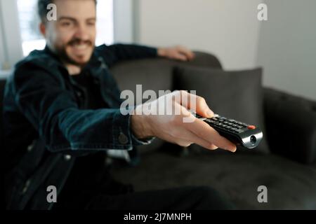 Happy young man wearing denim clothes with remote control switching channels on TV at home. Happy young guy watching interesting program on television Stock Photo