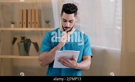 Hispanic physician male doctor man therapist medic wear uniform standing in hospital clinic writing notes fill medical form insurance notice detailed Stock Photo