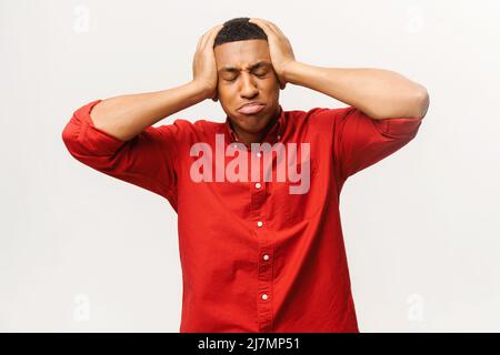 Upset young African-American short-cutted guy in red shirt holds head with both hands and suffers from strong headache isolated on white background, chagrin multiracial man in grief Stock Photo