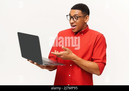 Amazed handsome African-American male entrepreneur in casual red shirt and stylish glasses looking at laptop screen isolated on white background, a guy surprised with offer Stock Photo
