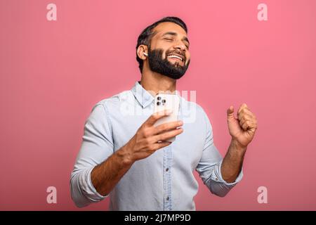 Excited satisfied multiracial man holding smartphone and rejoicing isolated on pink. Surprised emotional lucky Indian man with received good message, news, won the lottery Stock Photo