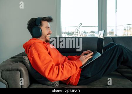 Handsome man laughing while using laptop with headset sitting on sofa in living room. Remote working or video call or watching video concepts. Stock Photo