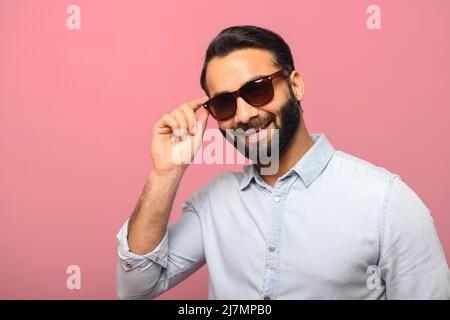 Headshot of handsome positive bearded man wearing sunglasses and blue casual shirt posing over pink background, looks at the camera, flirting Stock Photo