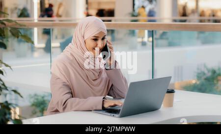 Arab multitasking woman sitting working on laptop talking on mobile telephone remotely consulting client using smartphone checks social network Stock Photo