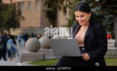 Young businesswoman freelancer student sitting outdoors reading email on laptop smiling laughing good news feels happy enjoys process surf web on Stock Photo