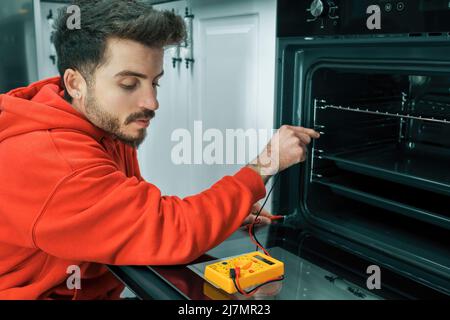 Young man wearing red hoodie with multimeter repairing oven, closeup. Stock Photo