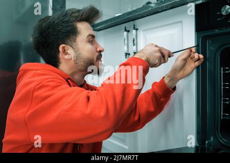 Young man wearing red hoodie repairing modern oven in kitchen with screwdriver. Stock Photo