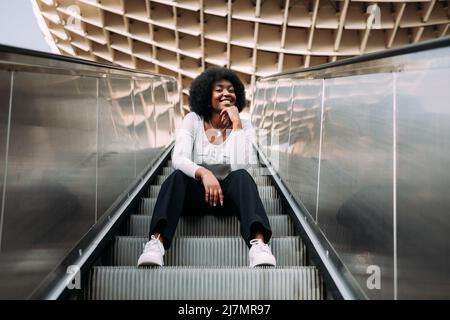 Young smiling black teenage woman with afro hair sitting on an outdoor escalator. Stock Photo