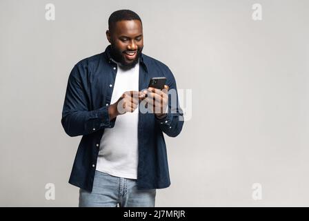 Happy African-American man in casual shirt over t-shirt using smartphone isolated on grey, black guy chatting online, using new mobile app, full length Stock Photo
