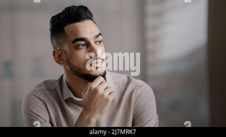 Thoughtful serious young hispanic man guy doubtful male professional worker sit at home office desk look away hold hand on chin lost in thoughts Stock Photo