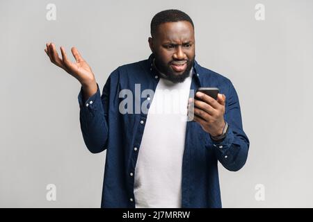 Disappointed young African-American man holding smartphone and watching at the phone screen, guy received bad notification or messages, feels upset, shrugging, isolated Stock Photo