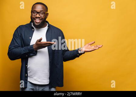 Smiling cheerful young African American man points both hands aside looking at the camera with cheerful wide toothy smile, black guy presenting novelty, advertising, isolated on gray wall Stock Photo