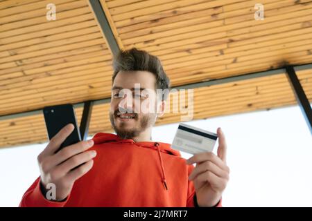 Happy young man looking phone screen and holding credit card for ordering products in internet. Fast online shopping. Stock Photo