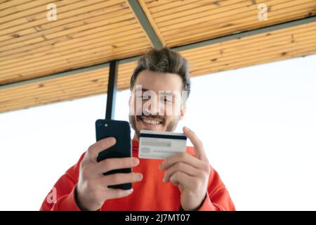 Fast online shopping. Happy young man using phone and credit card for ordering products in internet. Stock Photo