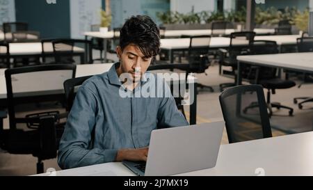 Young man Arabian employee writer thoughtfully typing laptop keyboard copying text in electronic form focused man checking original documents writing Stock Photo