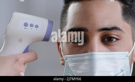 Close-up view infrared scanner on male customer's forehead arabian man in protective medical mask scanning body temperature patient measuring fever by Stock Photo