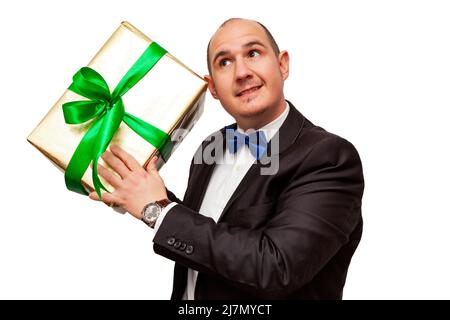 A smiling bald Caucasian adult male wearing a black suit, white shirt and blue bow tie is holding in his hands a gift wrapped with gold foil and a gre Stock Photo