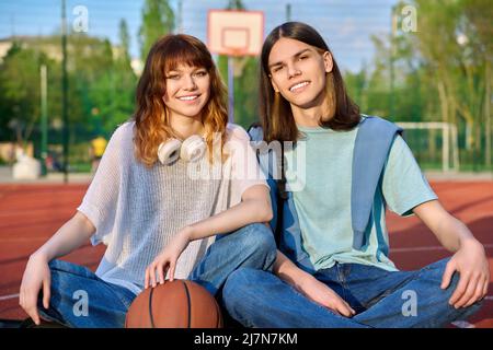 Friends teenagers guy and girl looking at camera, sitting on basketball court Stock Photo