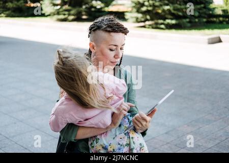 Freelance Parents. Busy working mom with baby toddler daughter on hands walk and looking on tablet in street. Multitasking Freelancer mom making notes Stock Photo