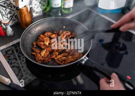 Woman cooking chicken teriyaki in frying pan on stovetop with spatula in home kitchen, steam rising during food preparation Stock Photo