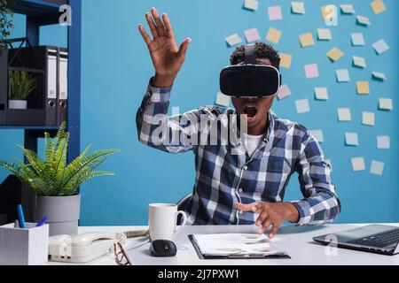 Astonished scared office worker wearing virtual reality headset while enjoying simulation game. Marketing agency amazed surprised employee playing with VR technology while in office space. Stock Photo