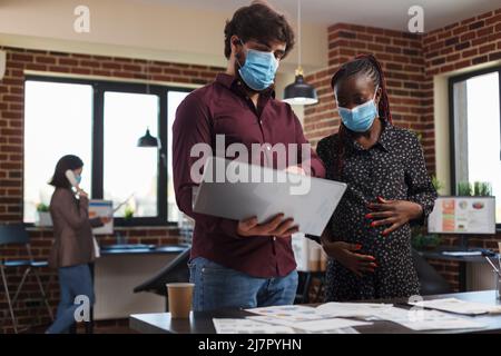 Multi ethnic diverse office workers wearing facemasks against coronavirus analyzing financial data and reviewing client portfolio. Expectant mother finance advisor looking over startup project ideas. Stock Photo