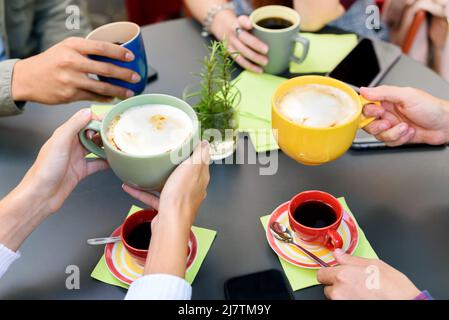 Crop unrecognizable group of friends drinking coffee and cappuccino from colorful cups while having breakfast together in light modern cafeteria Stock Photo