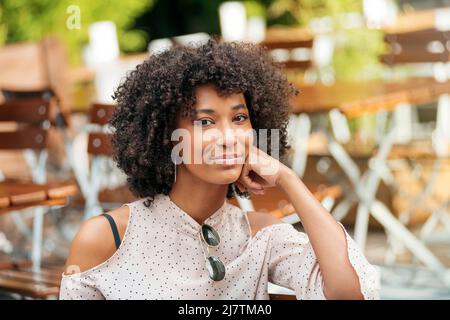 Attractive African American female with curly hair looking at camera while sitting on terrace of cafe with tables on blurred background Stock Photo