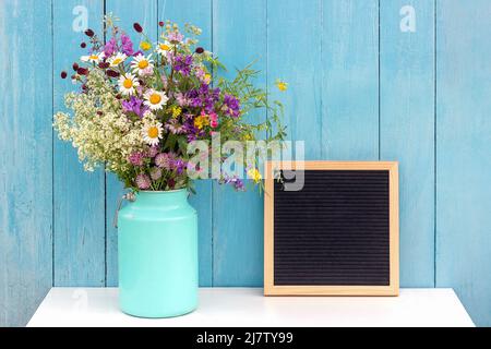 Holiday mock up. Empty blank black letter board and bouquet of bright flowers in tin can vase on table against blue wooden wall. Front view, Mockup. Stock Photo