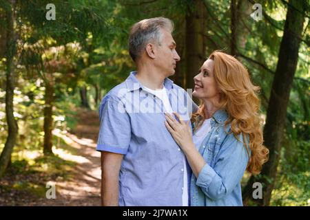 Smiling senior couple standing in park hugging each other together and smiling with happiness against the backdrop of trees on a warm day, enjoying retirement caucasian adult man and woman Stock Photo