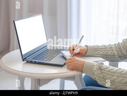 woman taking notes in notebook while using mockup laptop with blank white screen at home. freelancer writing details on book while working on laptop i Stock Photo