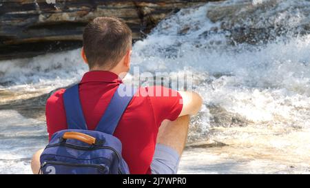 Back view of a male traveler in a red polo sitting by a mountain river Stock Photo