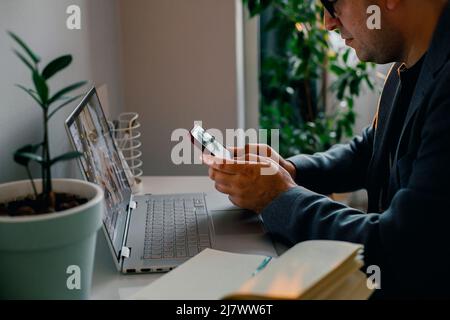 Focused European young man wearing glasses using laptop, texting on phone, working online on computer at home. Stock Photo
