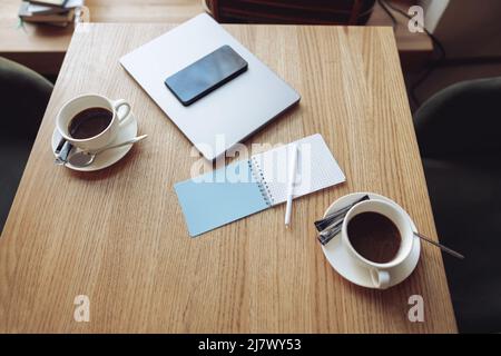 From above on table in cafe with coffee in two cups, laptop, smartphone and notebook with pen. Stock Photo