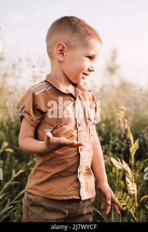 A little boy stands in the grass in nature and holds a snail in his hand Stock Photo