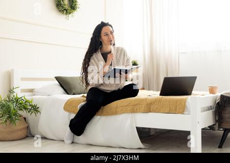 Woman sitting on bed, using computer and writing in notebook Stock Photo