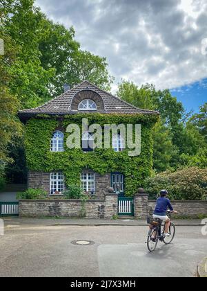 Essen, Germany – May 19, 2020: Beautiful overgrown vine leaf covered house in the district “Margarethenhöhe” with a woman on a bicycle in the foregrou Stock Photo