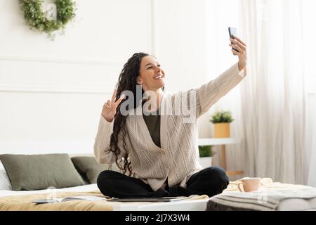 Young smiling woman sitting on bed taking selfie Stock Photo