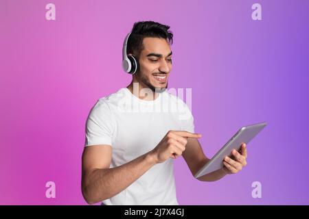 Positive young Arab man having video call, using tablet pc, wearing wireless headphones in neon light Stock Photo