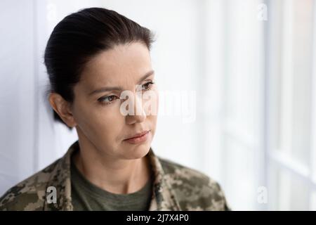 Thoughtful female soldier wearing camouflage uniform looking through window at home Stock Photo