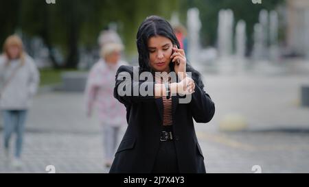 Young businesswoman excited calls on mobile phone angry that person late for meeting at scheduled time looking at wrist watch with irritation hispanic Stock Photo